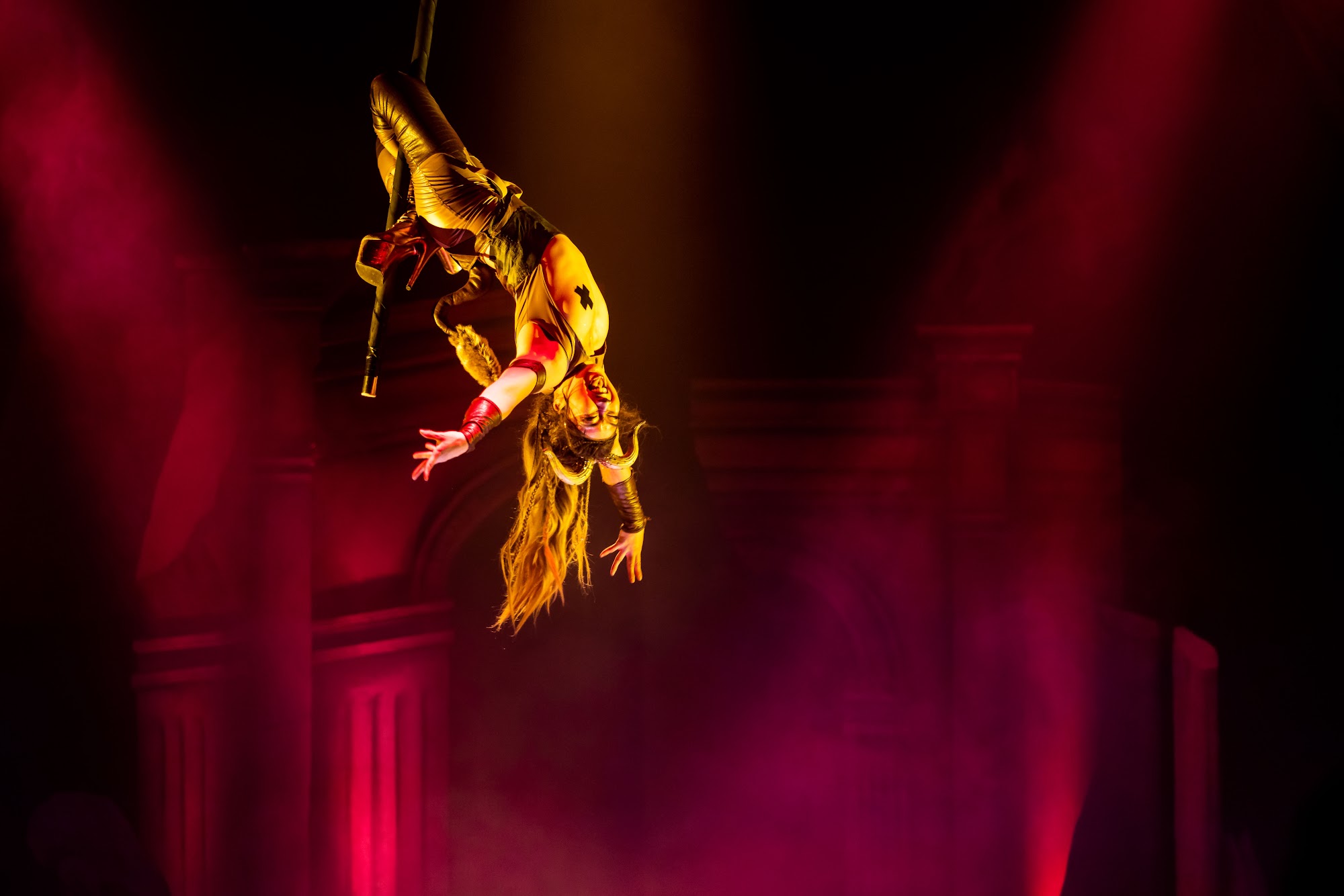 An inverted pole dancer hangs upside down in a crossed-leg layback on a flying pole, with arms extended and a large grin on her face.