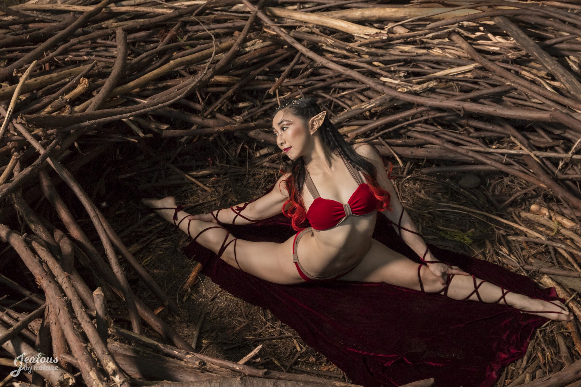 A pole dancer in a red bikini and elf ears does a split in a large bird's nest, surrounded by sticks.