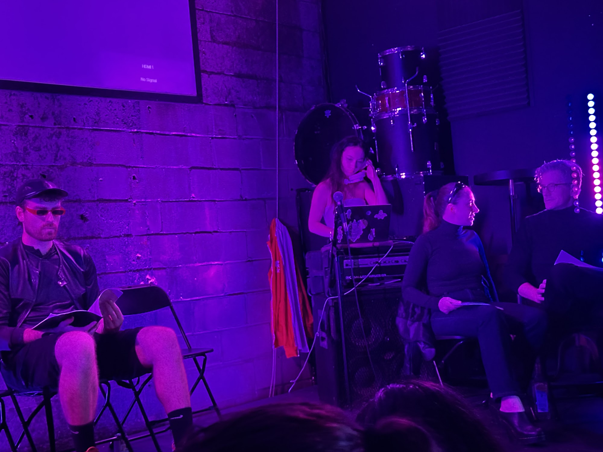 Three performers on stage during Meltdown at Unit J, Bushwick, 2026, with one in sunglasses reading from a script.