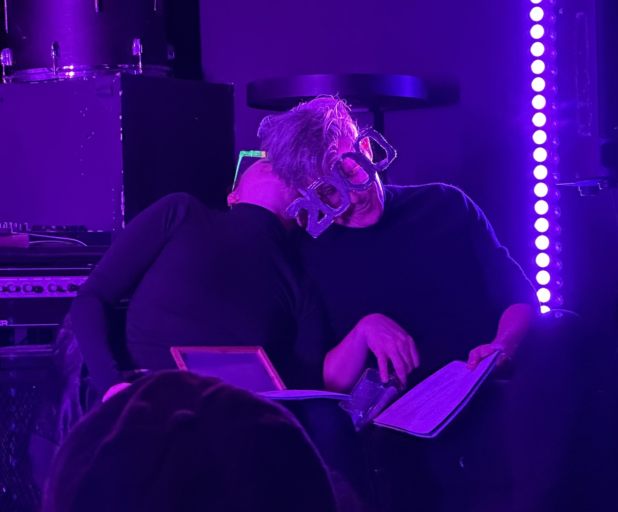 Two performers in black turtlenecks lean on each other, one wearing oversized glasses with 2000 on them.