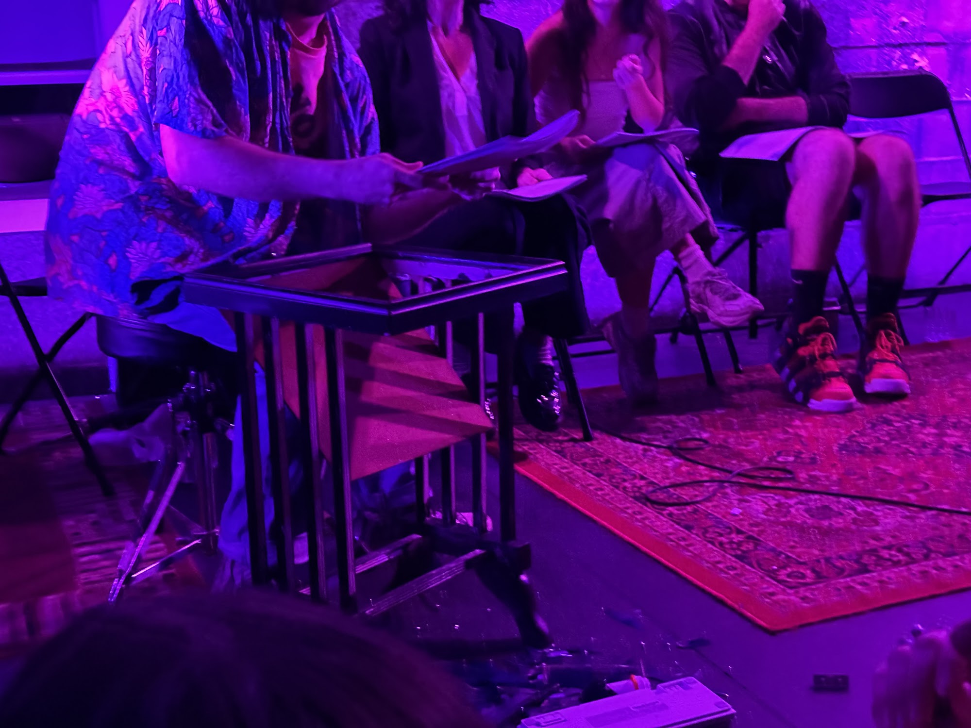 A performer in a colorful shirt leans over a small table on stage while others watch.