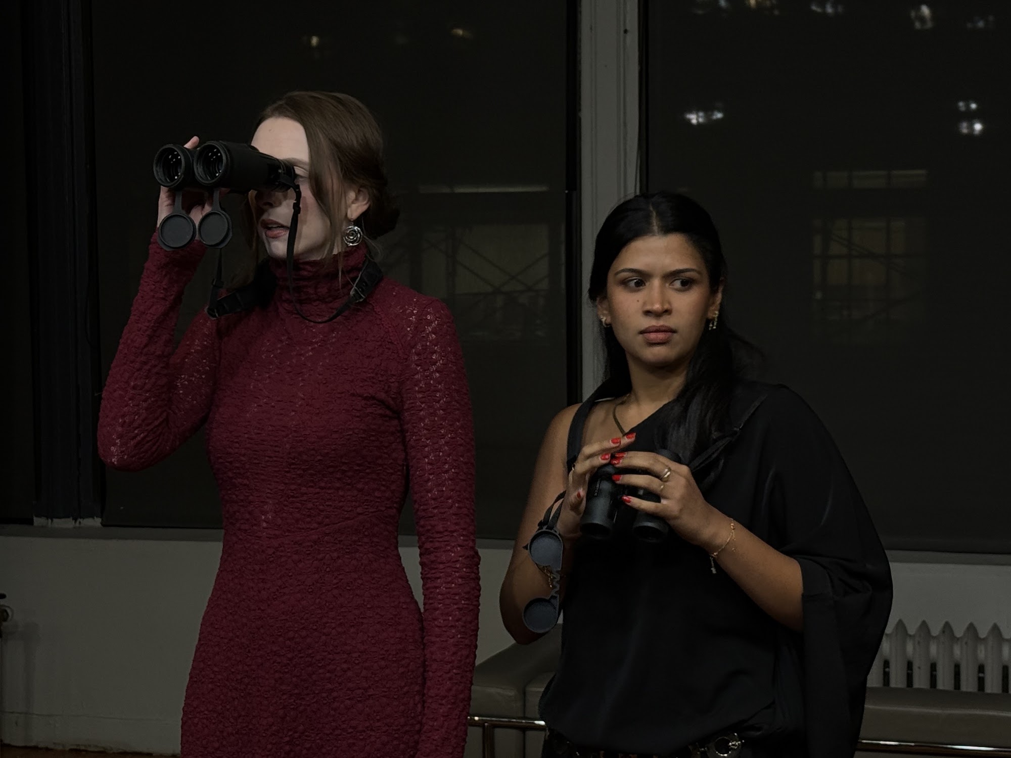 A woman in a black dress leans over another woman in a white dress while holding her face in a room.