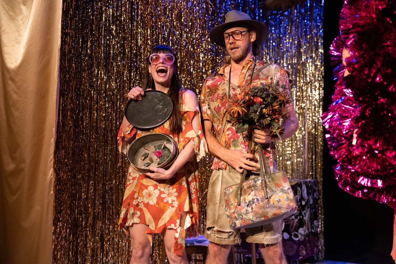 A performer in a dramatic costume poses on a stool, extending one leg and holding a microphone.