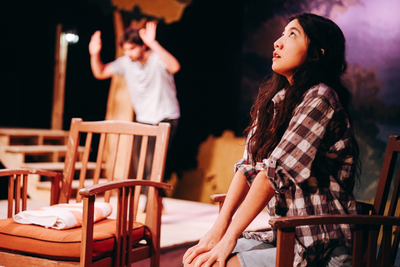 A woman sits on stage, looking upward, while a man behind her is raising his hands as if in celebration.