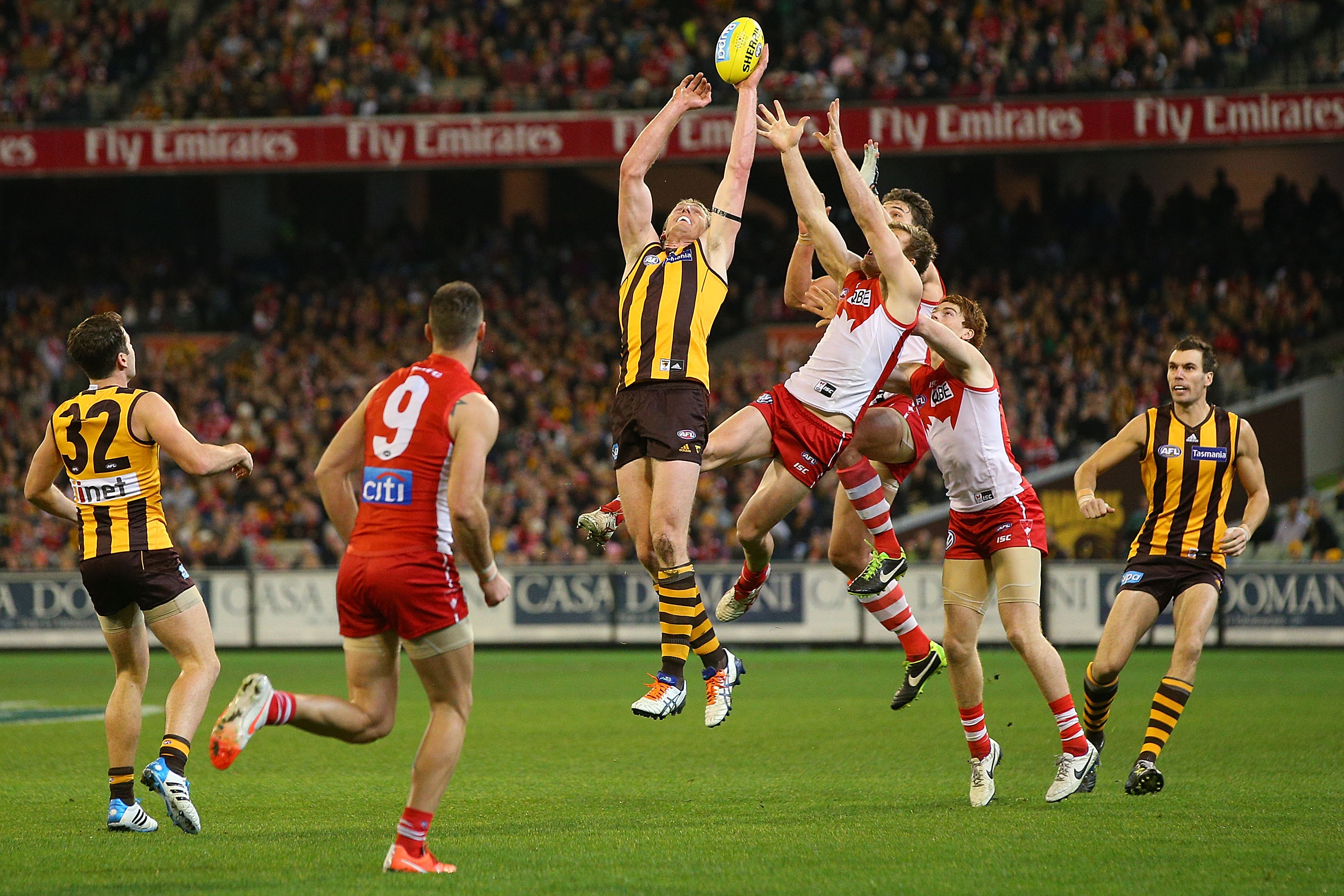 Australian rules football players compete to catch a ball mid-air during a match in a stadium.