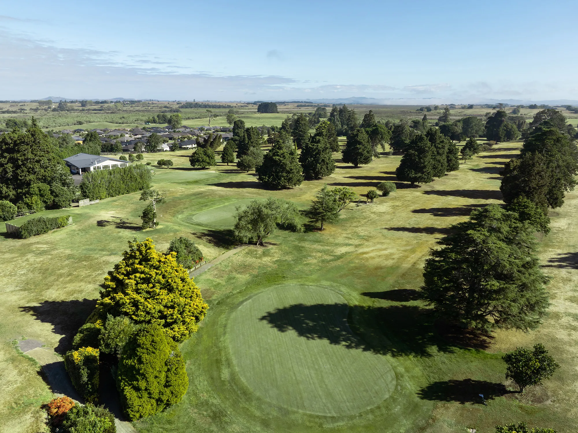 Aerial view of a golf course with green fairways, scattered trees, a clubhouse, and a residential area in the background under a partly cloudy sky.
