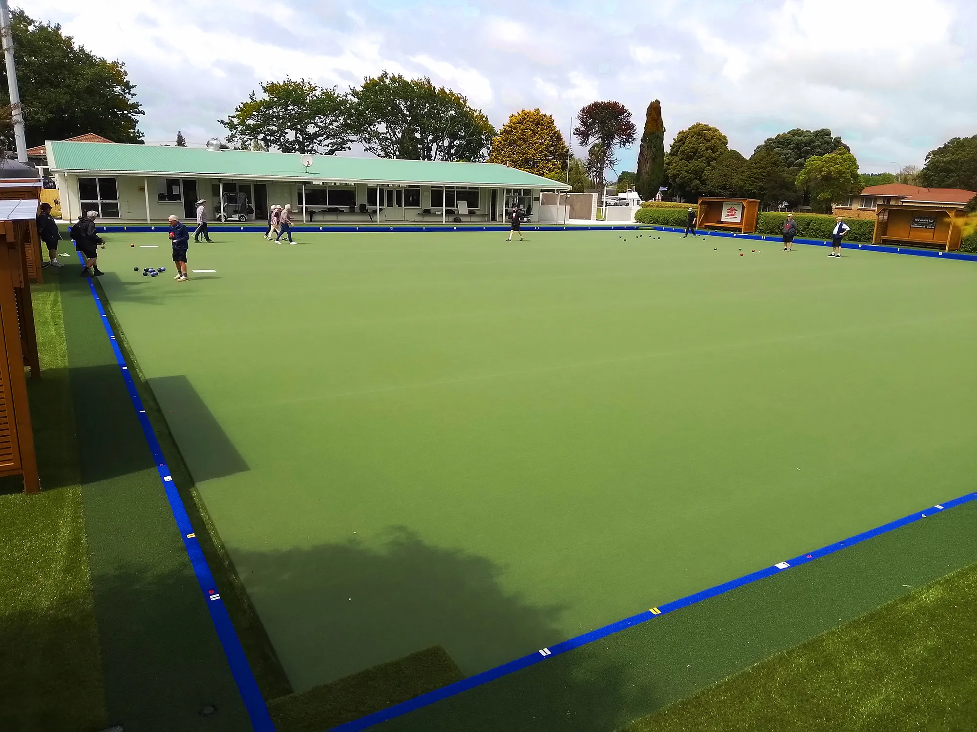 Group of people playing lawn bowls on a green bowling lawn at Te Kauwhata Bowling Club with a building and trees in the background.