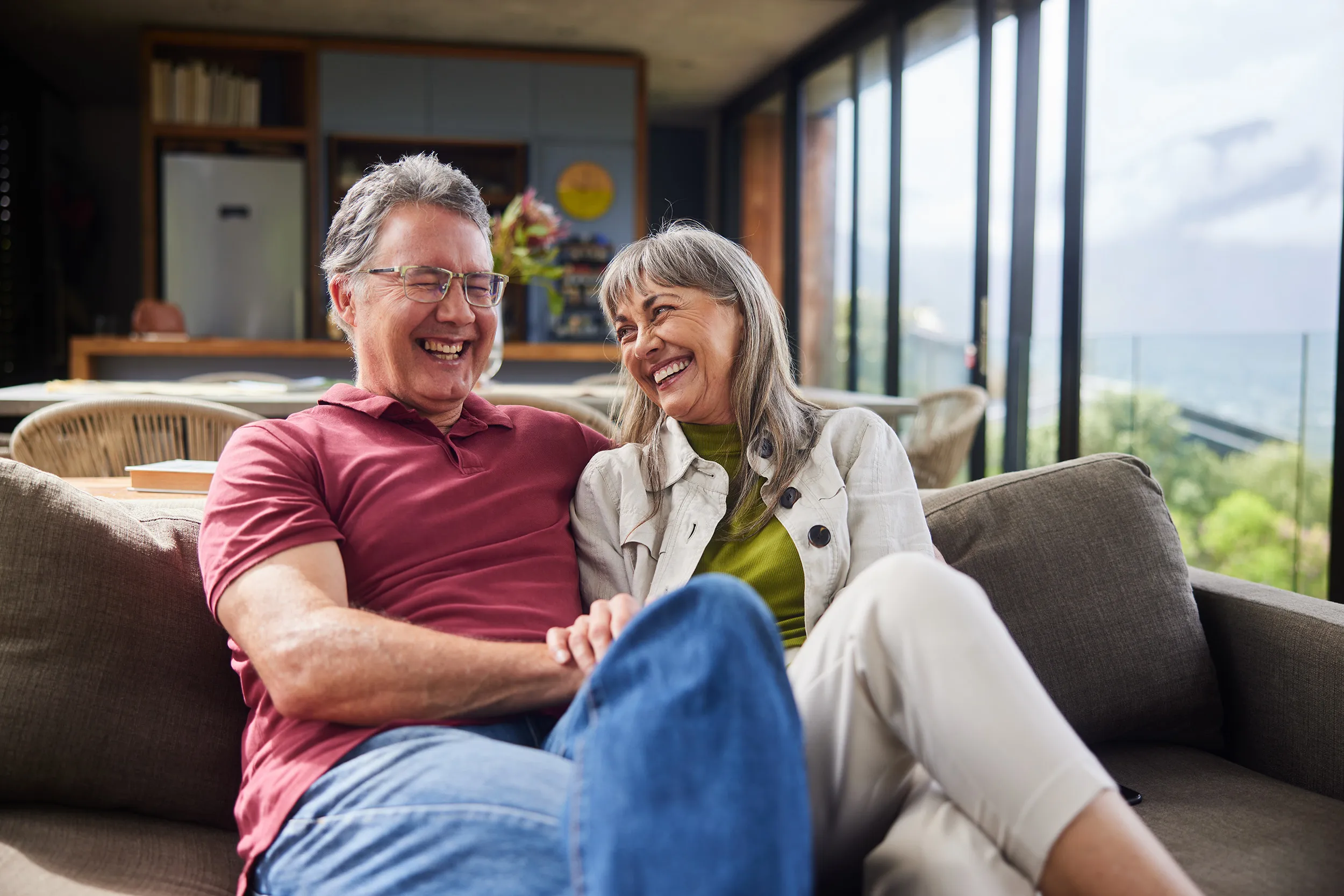 Smiling older couple sitting together on a coach in a bright room with large windows overlooking greenery and water.