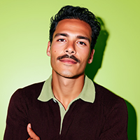 Young man with a mustache wearing a dark sweater over a green collared shirt against a green background.