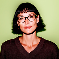 Portrait of a woman with short dark hair and bangs wearing large round glasses and a dark brown V-neck sweater against a light green background.