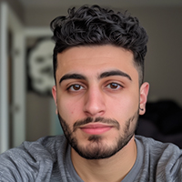 Young man with curly black hair, trimmed beard, and gray sweater looking directly at the camera indoors.