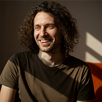 Man with curly hair smiling while sitting in warm sunlight wearing a dark shirt.