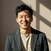 Smiling man with dark hair wearing a gray blazer and beige shirt in sunlight with shadows on the wall.