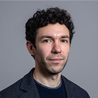 Portrait of a man with curly dark hair wearing a dark blazer and shirt against a gray background.