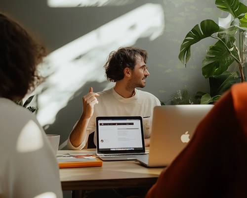 Man sitting at a table with laptops, pointing to the side while in a discussion with two other people.