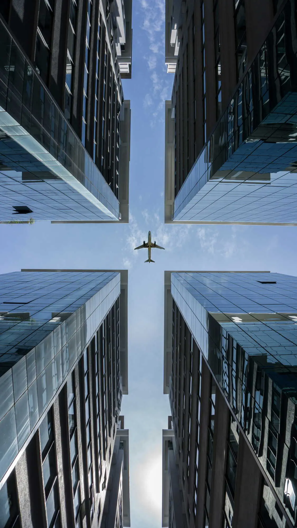 Photographie prise du sol montrant un avion volant entre quatre gratte-ciels en verre reflétant le ciel bleu.