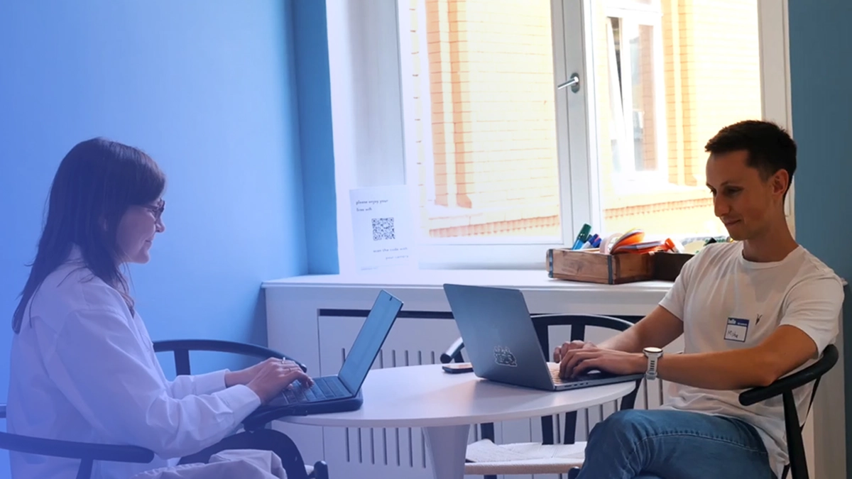 Two people sitting at a round table in a bright room, each working on a laptop.