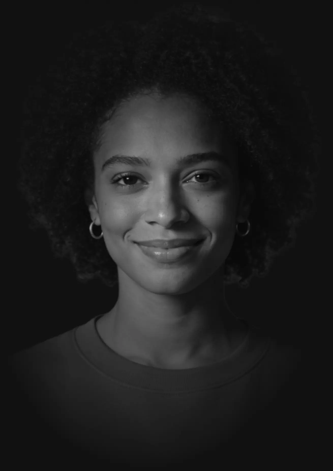 Black and white portrait of a smiling woman with curly hair and hoop earrings against a dark background.