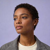 Portrait of a thoughtful young woman with short curly hair wearing a gray jacket and white shirt against a lavender background.