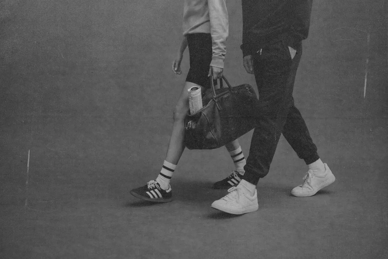 Black and white lifestyle photo of two people walking side by side in casual sportswear and sneakers. One person carries a leather gym bag with a water bottle inside. Minimal studio background, active urban fitness style, movement, wellness and everyday routine concept.