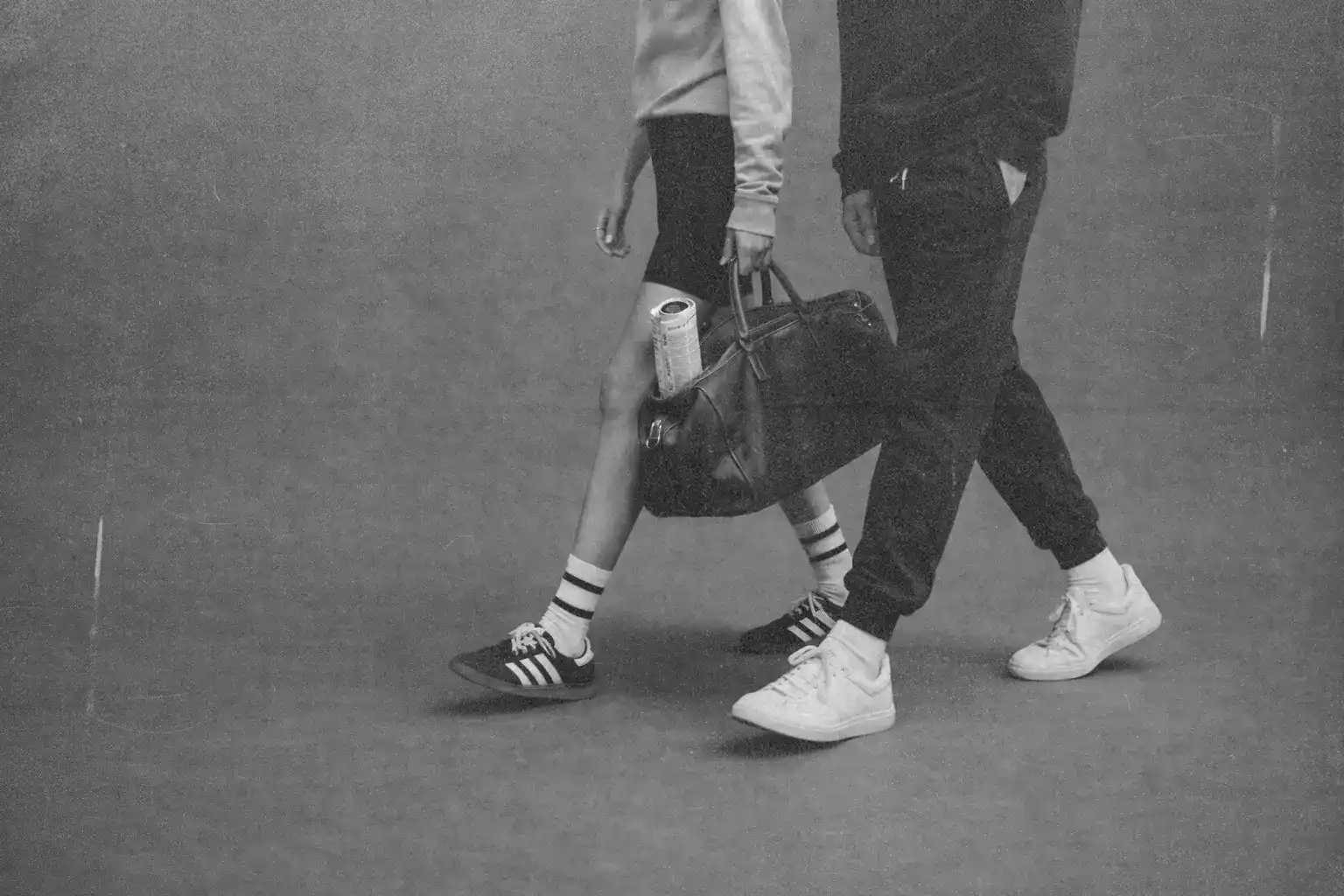 Black and white lifestyle photo of two people walking side by side in casual sportswear and sneakers. One person carries a leather gym bag with a water bottle inside. Minimal studio background, active urban fitness style, movement, wellness and everyday routine concept.