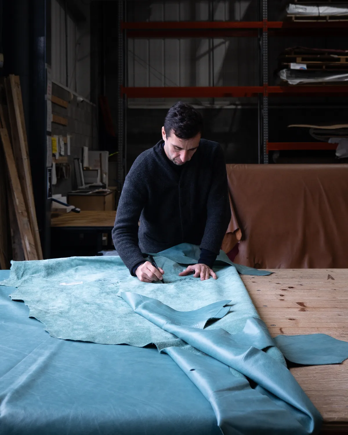 Man in dark sweater marking large teal leather fabric on a wooden table in workshop.