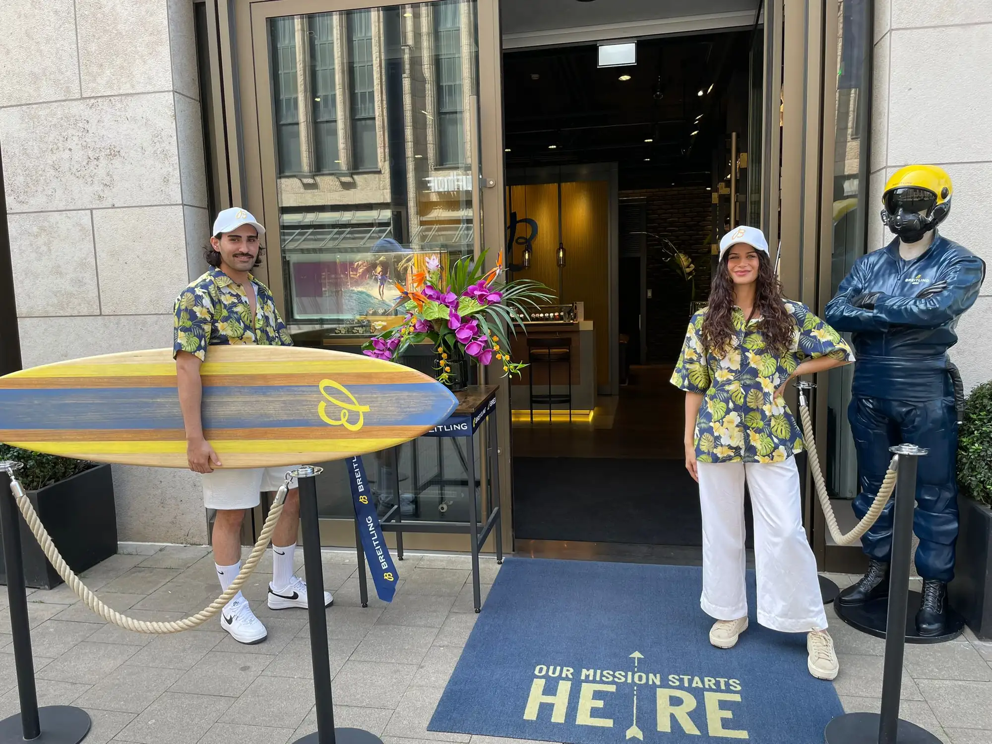 Zwei Hosts in Hawaii-Hemden und weißen Breitling-Caps mit Surfboard vor der Breitling Boutique Düsseldorf