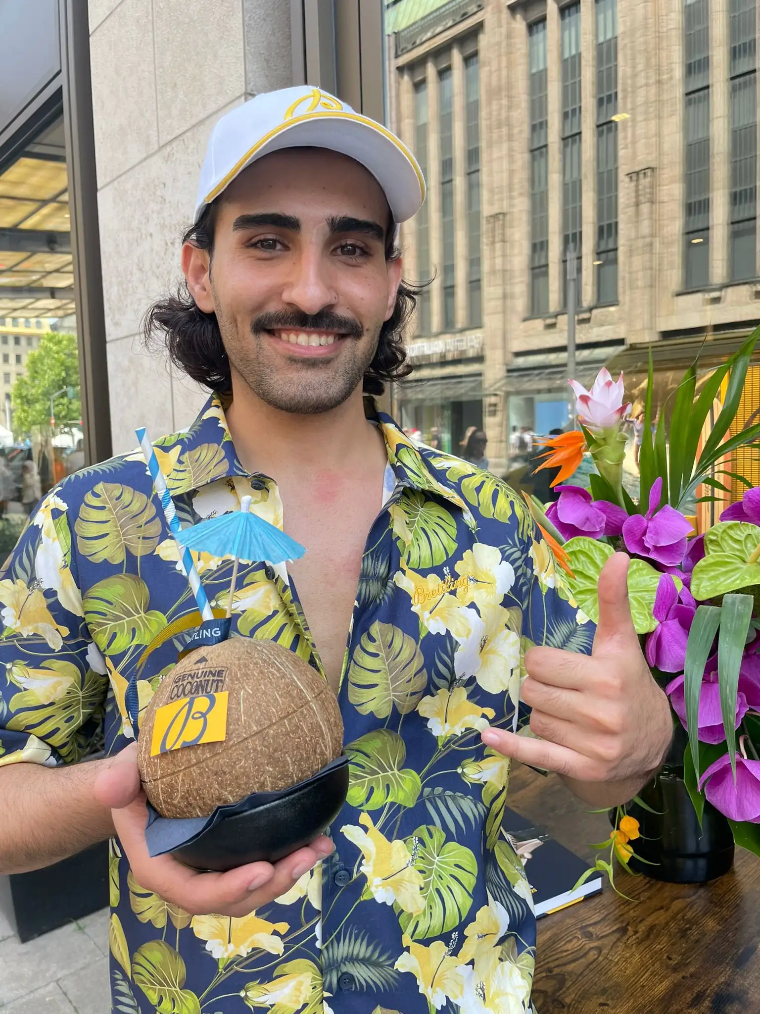 Host in a Hawaiian shirt and white Breitling cap holding a coconut drink with a blue paper umbrella outside the Breitling Boutique Düsseldorf