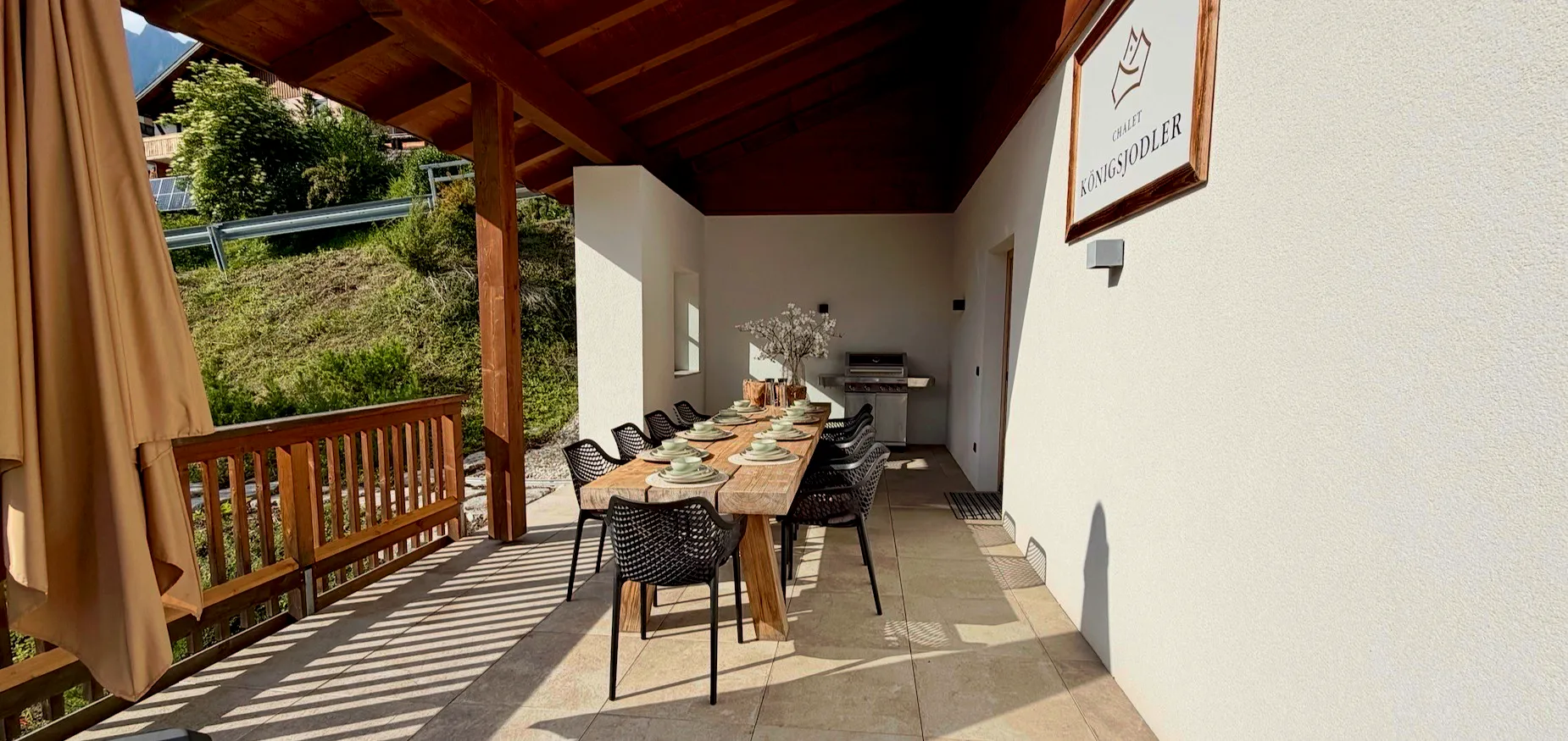 Outdoor dining area with a wooden table, black chairs, tableware, and a grill under a wooden roof.