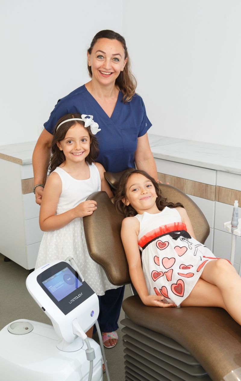 Medic dentist tânăr îmbrăcat în halat albastru, cu brațele încrucișate, în cabinet stomatologic cu scaun verde și echipamente dentare albe.