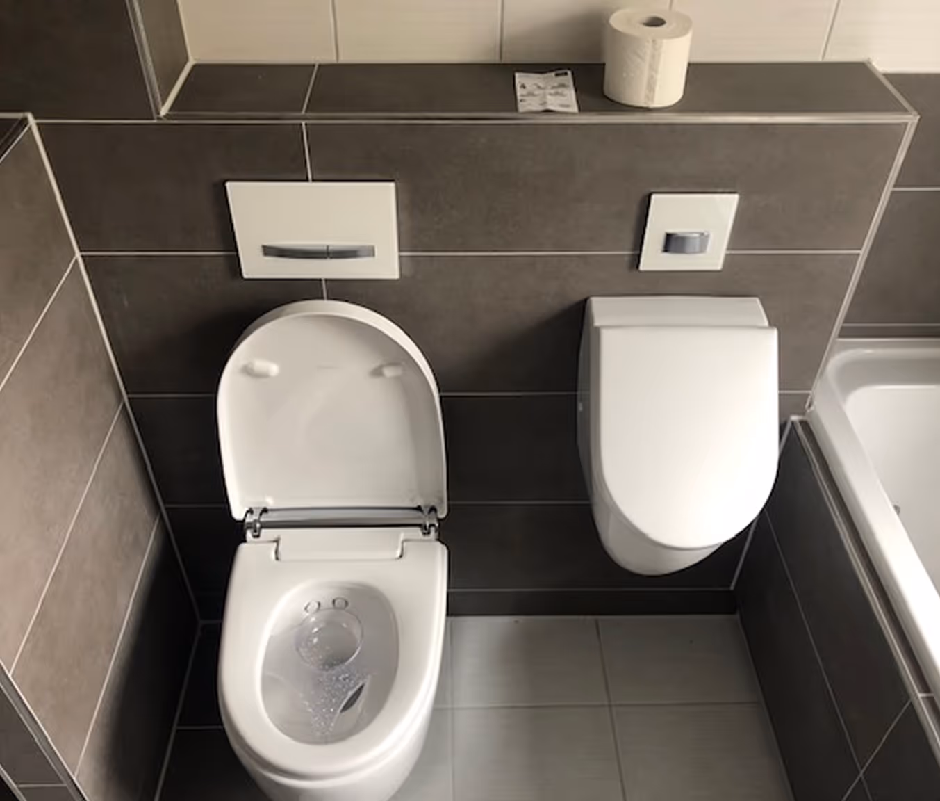 Moderne Toilette mit geöffnetem Sitz und geschlossenem Urinal an gefliester Wand, darüber eine Rolle Toilettenpapier.