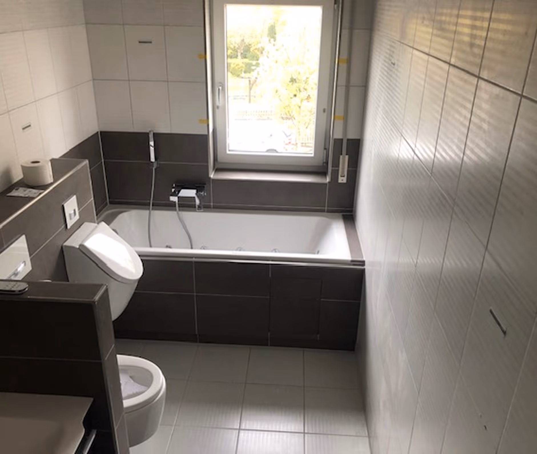 Modernes Badezimmer mit Badewanne unter einem Fenster, Toilette und grauen Fliesen.