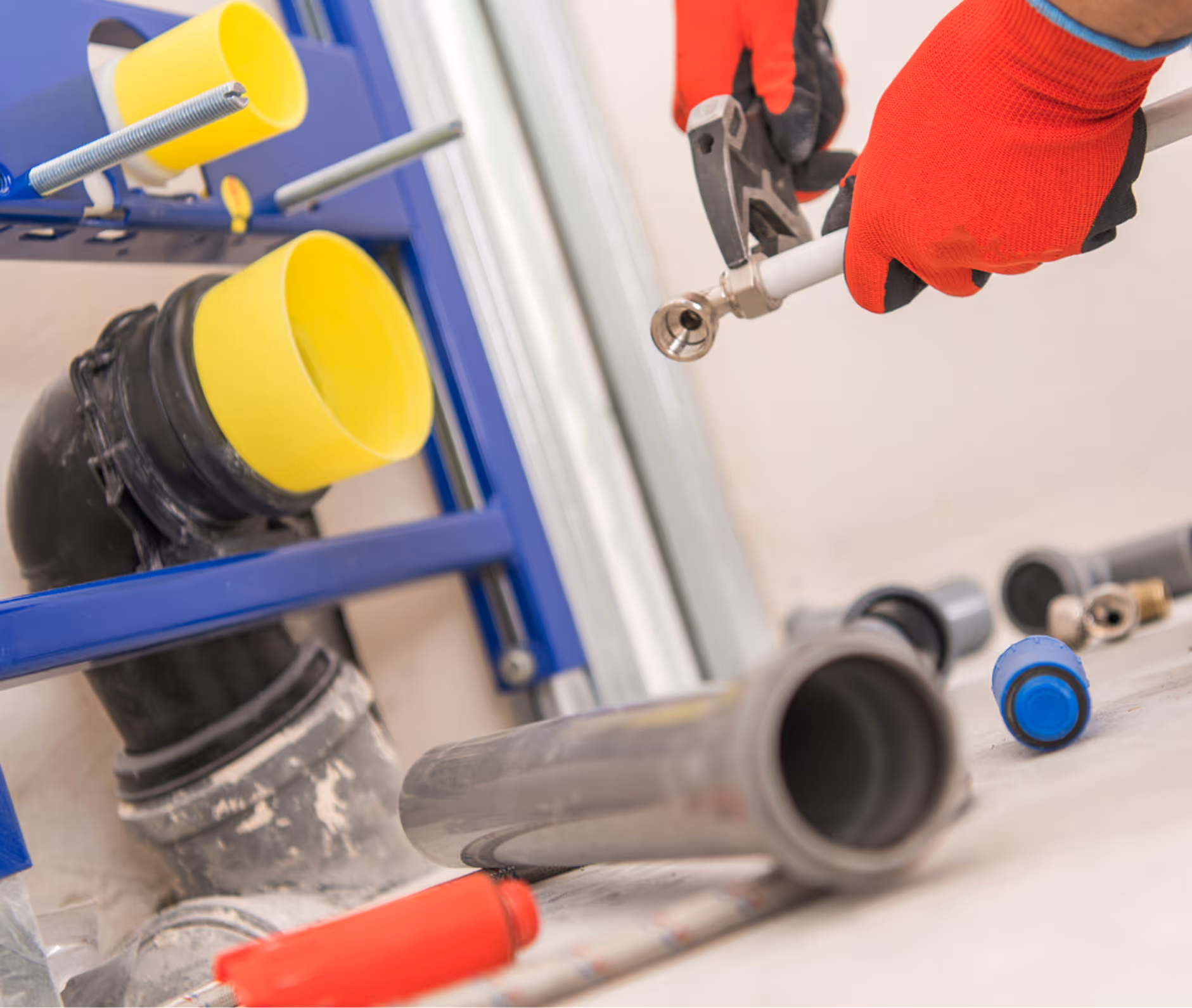 Hand in roten Arbeitshandschuhen hält eine Rohrzange an einem weißen Rohr, umgeben von verschiedenen Installationsrohren und Bauteilen.