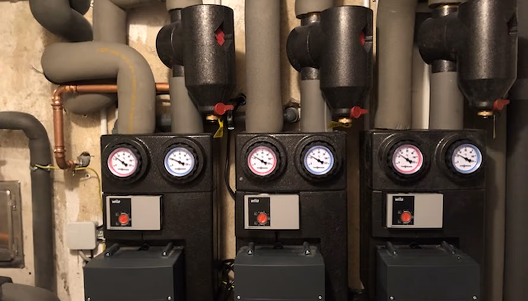 Drei Heizsystemeinheiten mit Druck- und Temperatursensoren in einem Kellerraum, umgeben von isolierten Rohren.