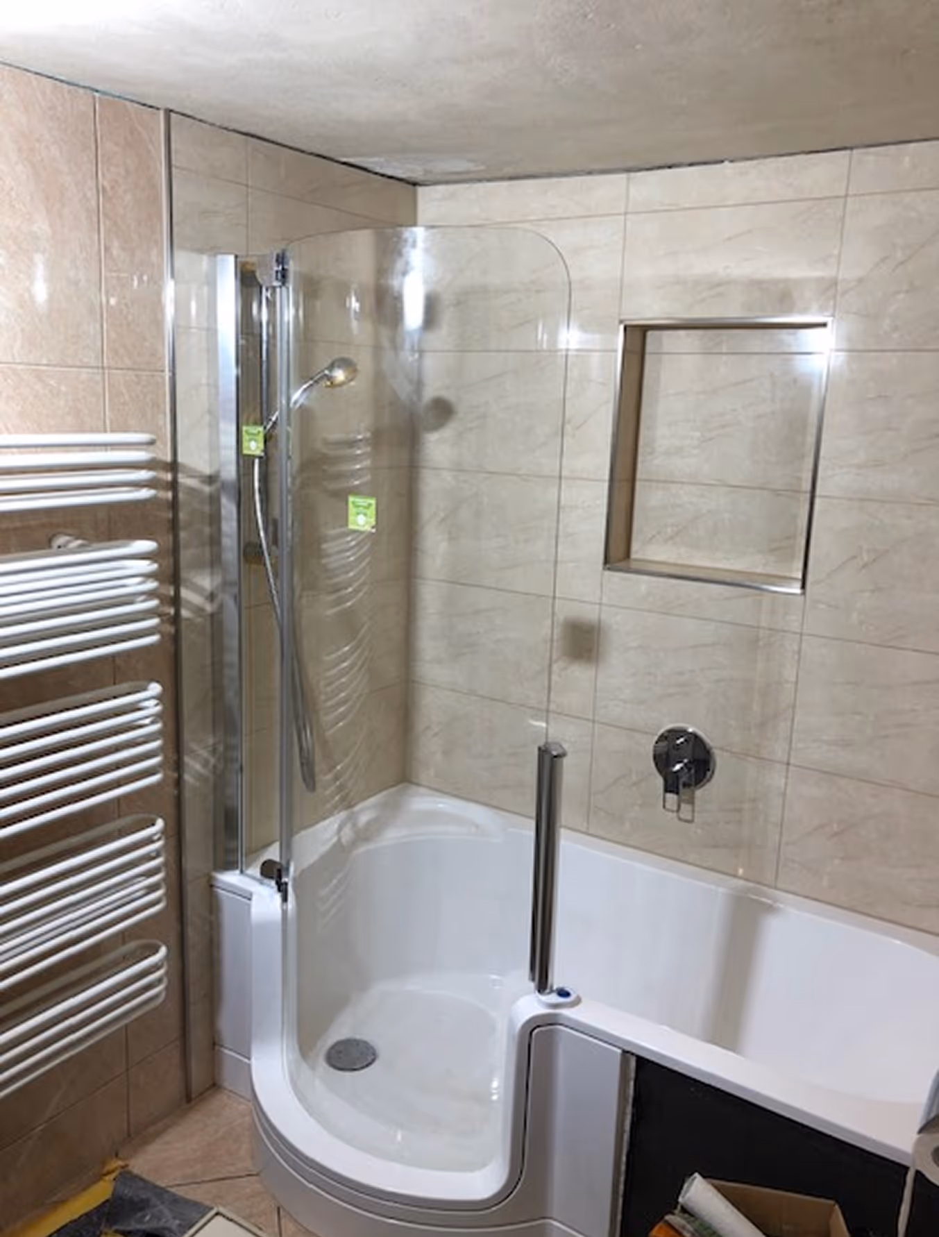 Barrierefreie Dusche mit halbkreisförmiger Einstiegstür, Duschkopf und Handtuchheizkörper in einem hell gefliesten Badezimmer.