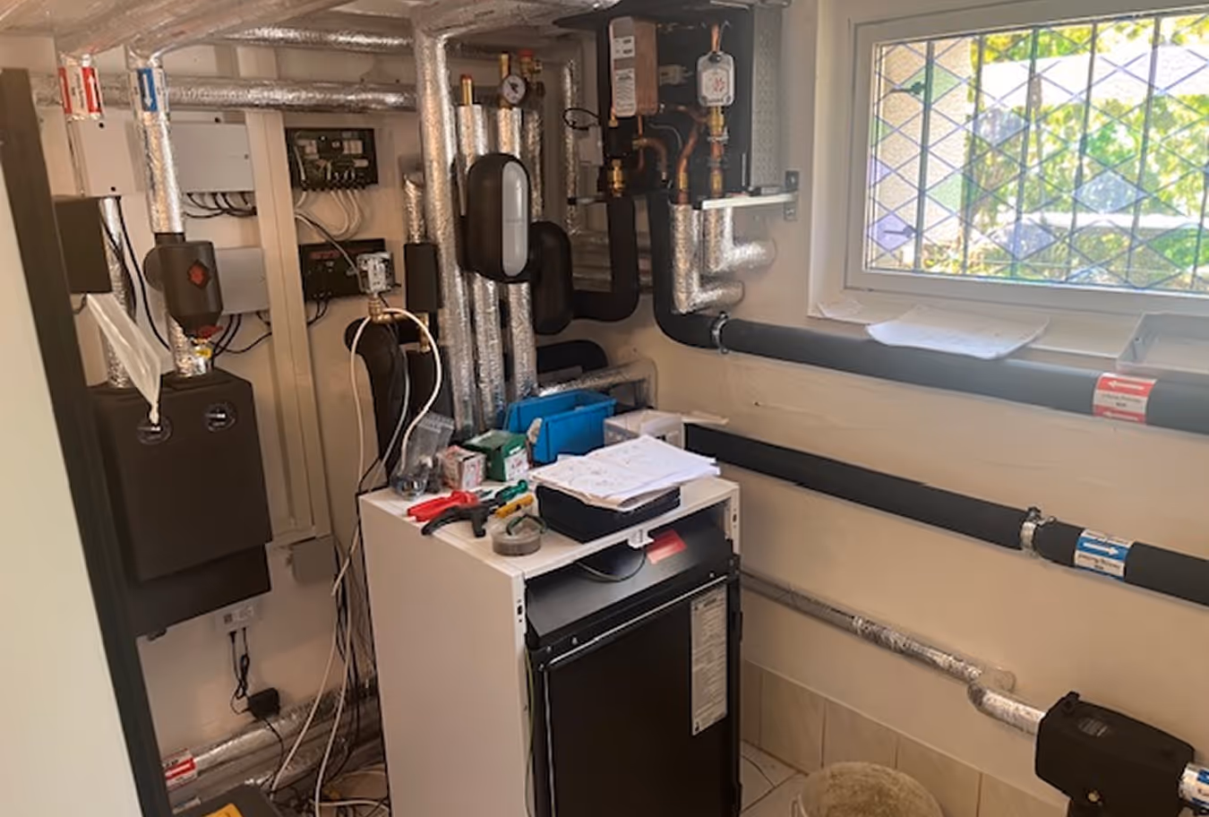 Heizungsraum mit isolierten Rohren, Steuergeräten und Werkzeugen auf einem weißen Schrank unter einem Gitterfenster.