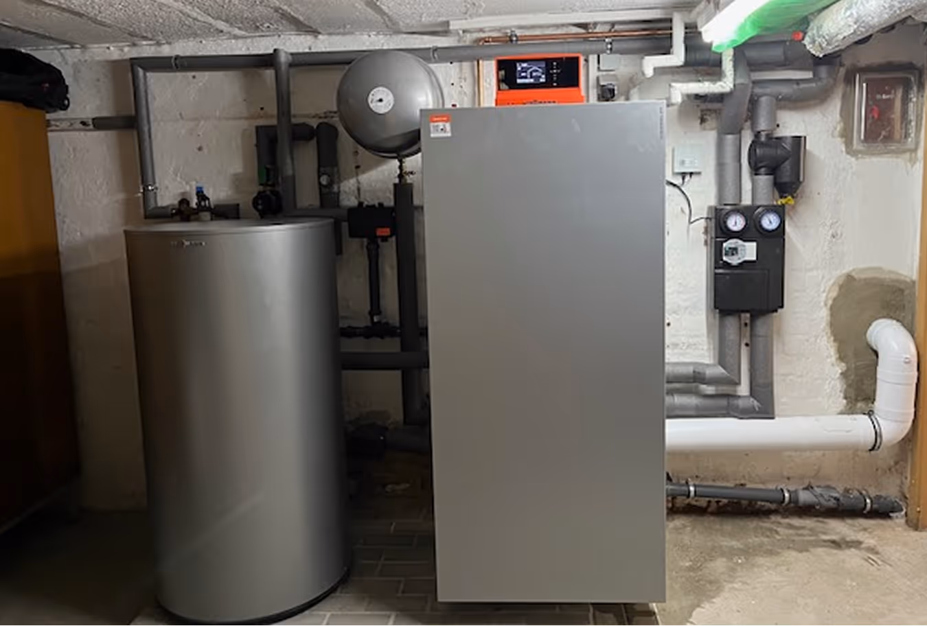 Heizungsanlage im Keller mit großem grauem Warmwasserspeicher und Heizgerät mit Rohren und Steuergerät an der Wand.