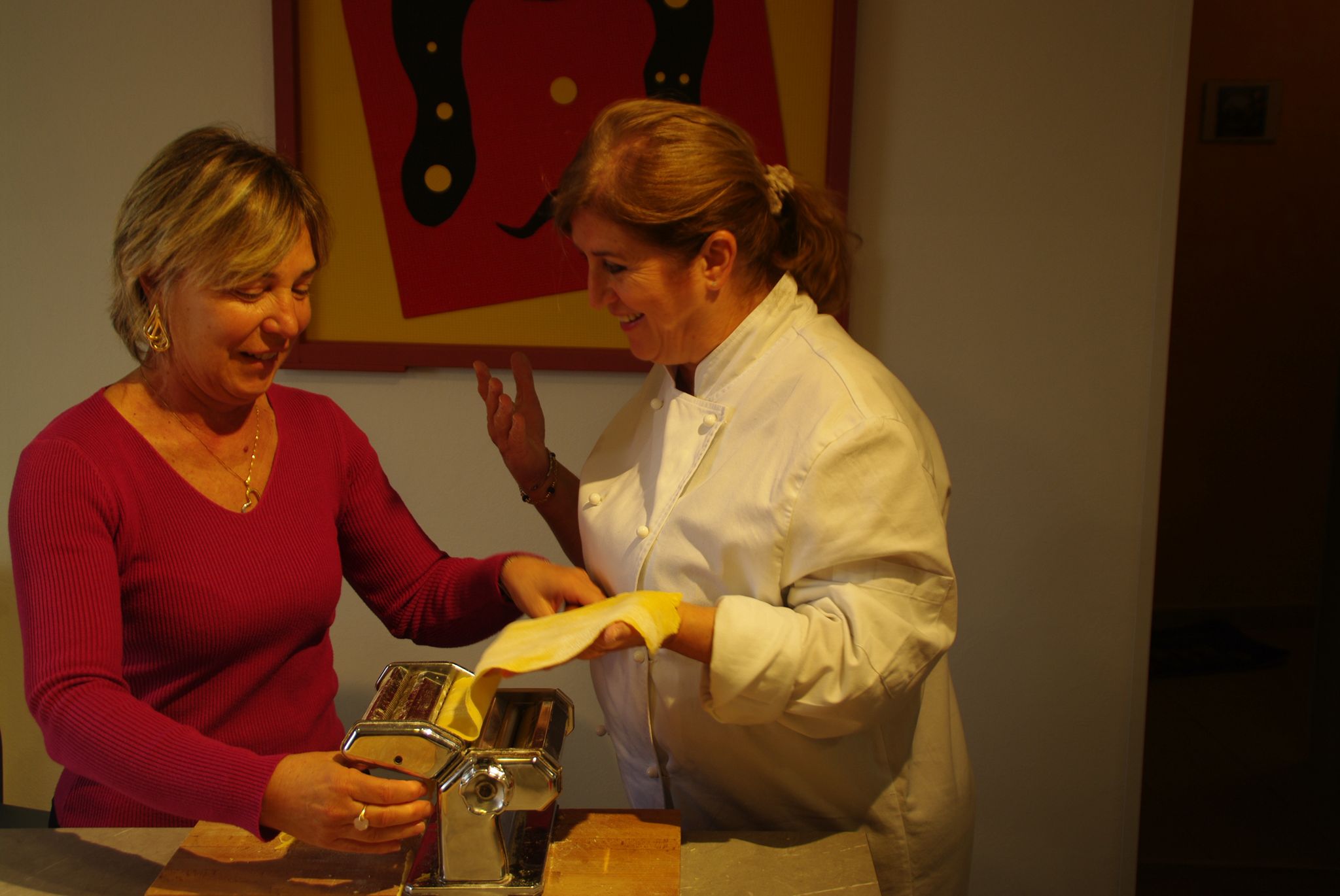 Two women making fresh pasta with a pasta machine, one feeding dough into the machine while the other turns the handle.