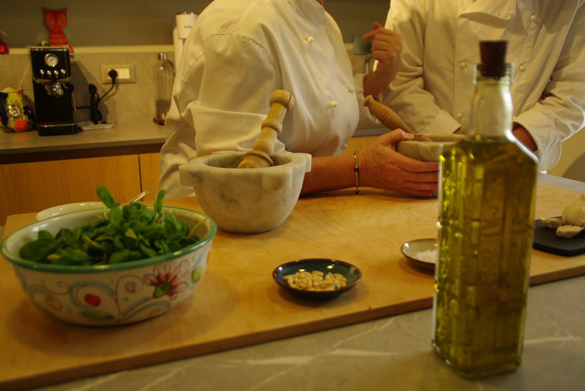 Two chefs in white uniforms using marble mortar and pestle on kitchen counter with a bowl of greens, pine nuts, and olive oil bottle nearby.