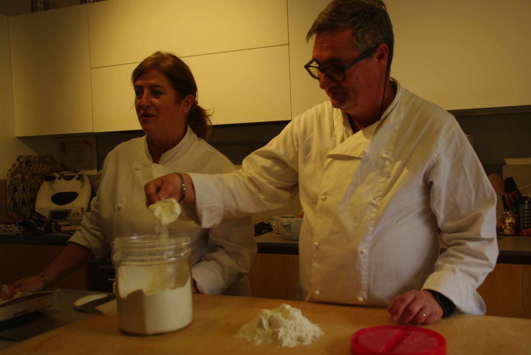 Two chefs in white coats preparing flour in a kitchen.