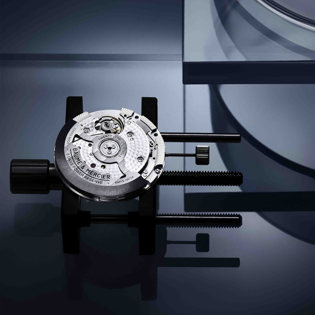 Close-up of Baume & Mercier Swiss watch movement in a holder on a reflective surface.