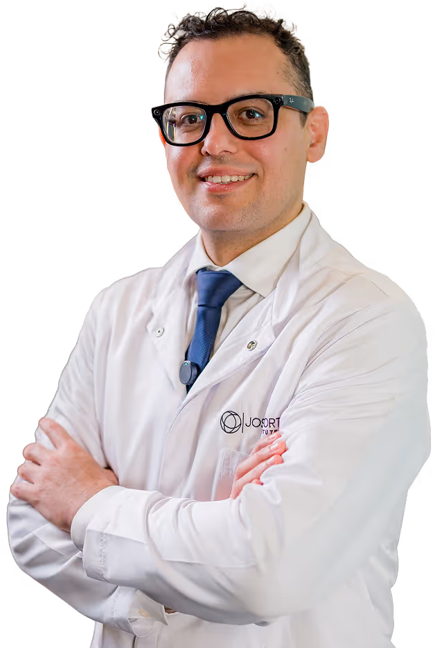 Man wearing glasses, a white lab coat, and a blue tie, standing with arms crossed and smiling.