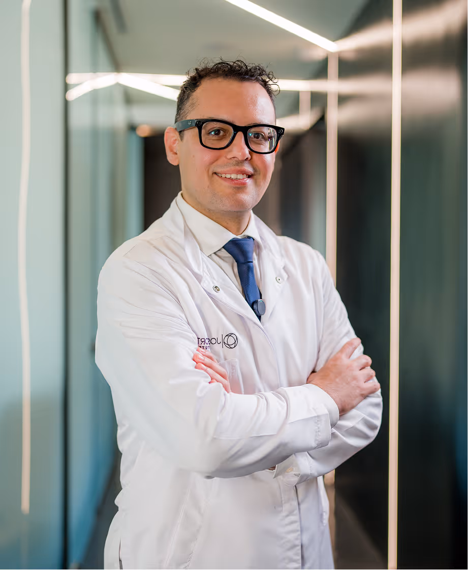 Smiling male doctor wearing glasses and a white lab coat with arms crossed in a modern hallway.