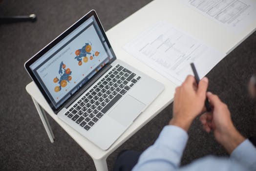 Professional analyzing data infographics on a laptop at an office desk, featuring documents and a pen.
