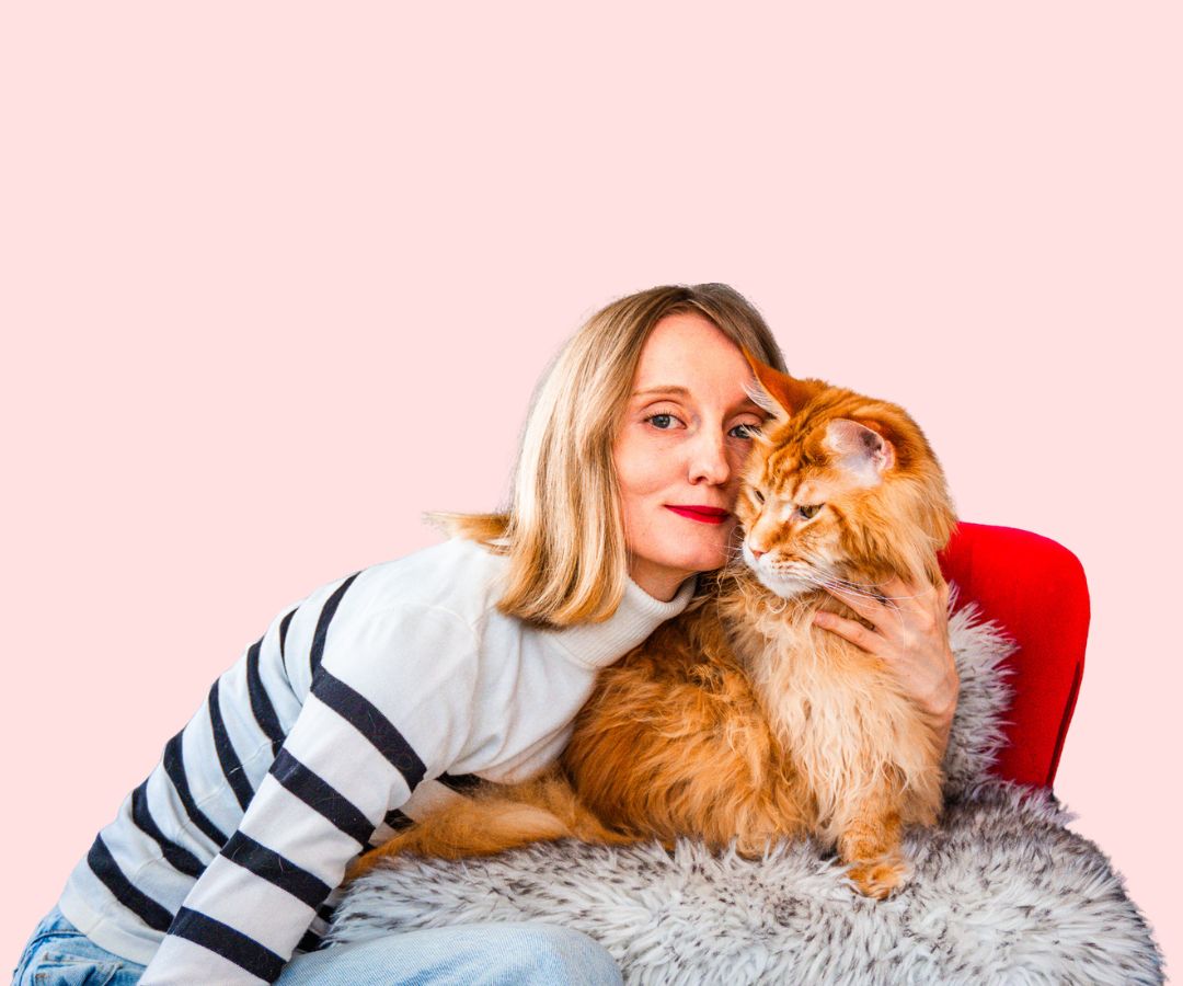 Woman with blonde hair hugging a large orange Maine Coon cat on a furry blanket and red chair.