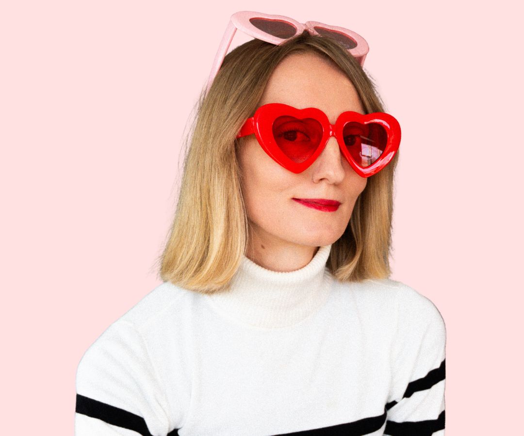 Woman with shoulder-length blonde hair wearing red heart-shaped sunglasses and a white turtleneck sweater.