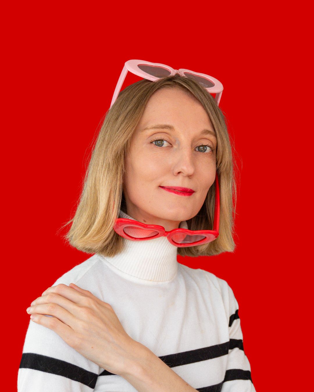 Woman with blonde bob wearing a white and black striped turtleneck and heart-shaped sunglasses on red background.