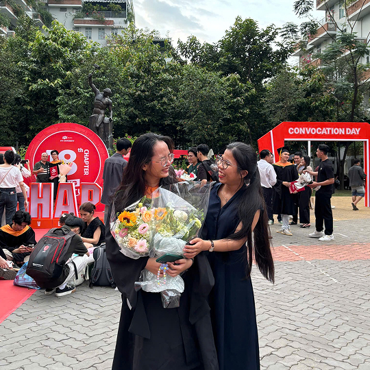 Two women smiling and embracing during an outdoor convocation ceremony, with one holding a bouquet of flowers.