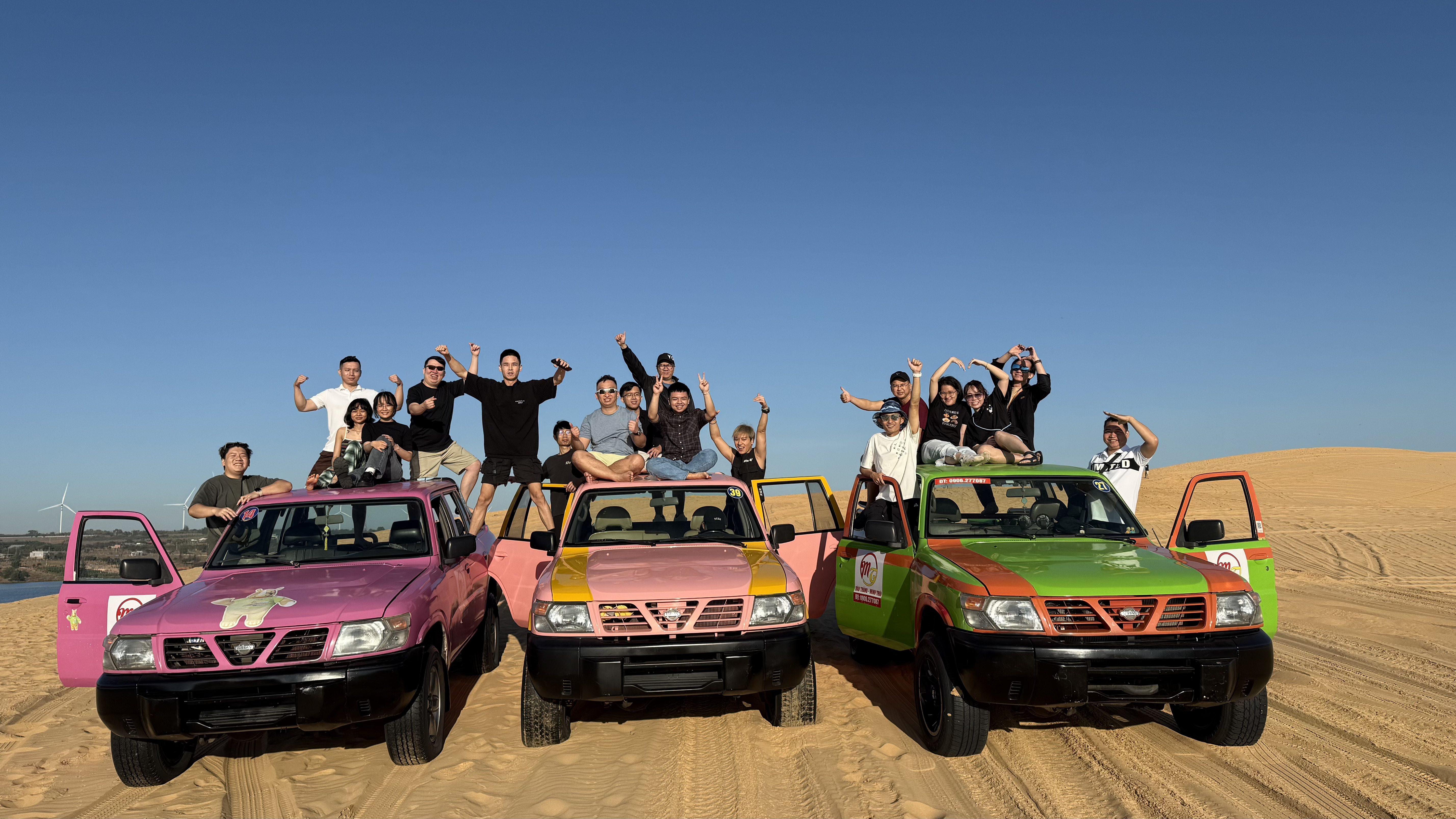 Group of people posing atop three colorful off-road vehicles on sand dunes under clear blue sky.
