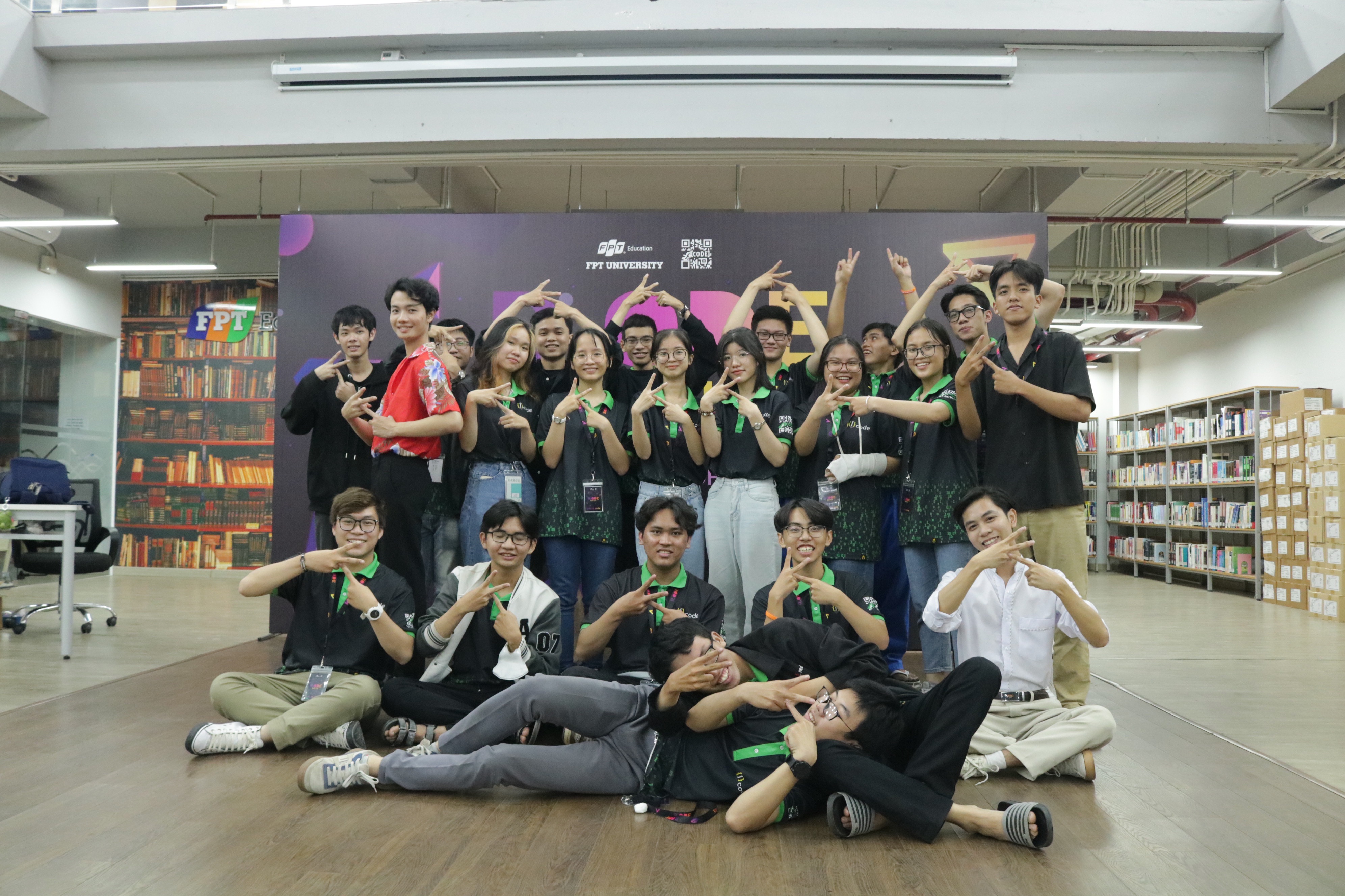 Group of young people posing indoors making hand gestures, with a backdrop displaying FPT University logo.